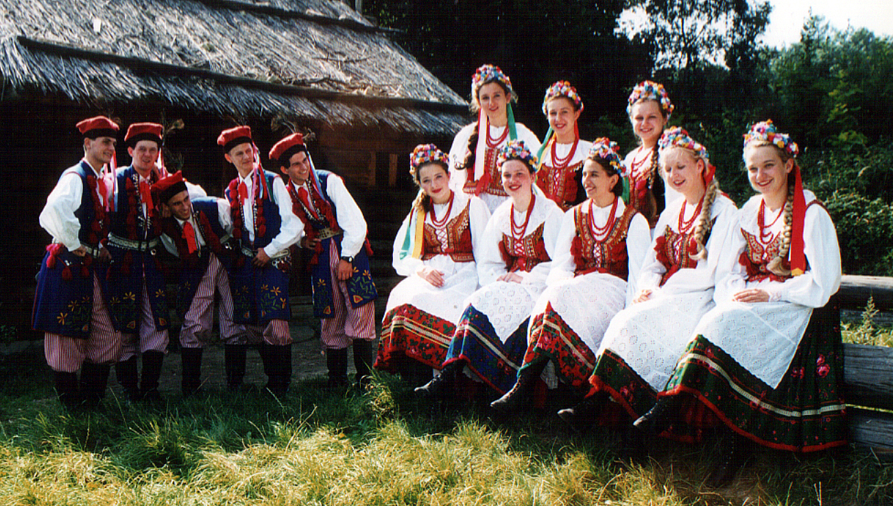 Foto von 2 Gruppen an Personen in traditioneller Kleidung vor einer Reedgedeckten Scheune/Haus.