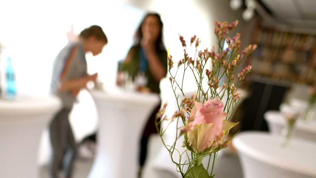 im Hintergrund sind zwei Frauen an einem Stehtisch zu sehen, im Vordergrund steht eine Vase mit einer Rose und Schleierkraut