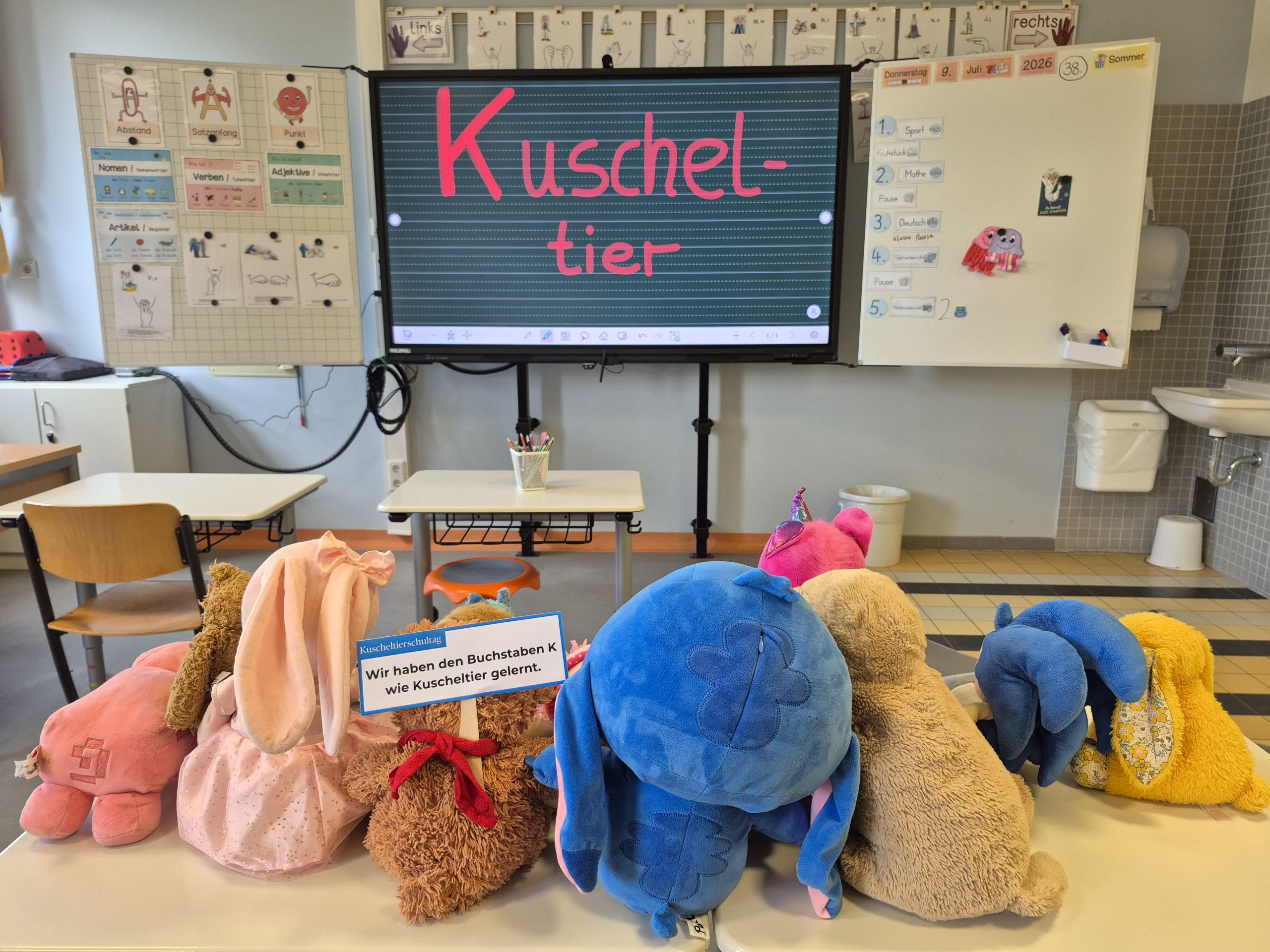 20260715_093758 Kuscheltiere sitzen vor einem Smartboard. Das Wort Kuscheltiere ist auf der Tafel geschrieben.