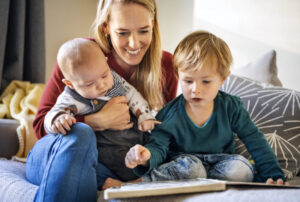 Eine lächelnde Mutter hält ihr Baby im Arm, während ihr Kleinkind auf das Buch zeigt; gemeinsam sitzen sie zu Hause und lesen.